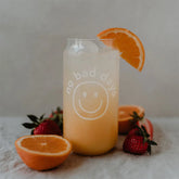 Ein Glas Orangengetränk mit Eis und einer Orangenscheibe im Eulenschnitt Hohes Trinkglas "No Bad Days", mit einem Smiley-Gesicht. Erdbeeren und Orangenscheiben schmücken die helle Fläche rund um das Glas.