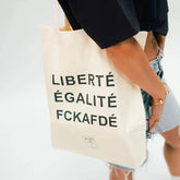 Eine Person in schwarzem Hemd und Jeans-Shorts hält die Arrell FCKAFDÉ Premium Tote Bag - Nature & Black in der Hand, die aus fair gehandelter Bio-Baumwolle besteht und mit "LIBERTÉ ÉGALITÉ FCKAFDÉ" in fetten schwarzen Buchstaben bedruckt ist.