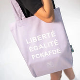 Jemand trägt die Arrell FCKAFDÉ Premium Tote Bag - Purple & Vanilla Ice, mit weißem "LIBERTÉ ÉGALITÉ FCKAFDÉ"-Schriftzug aus Bio-Baumwolle auf einfarbigem Hintergrund.