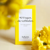 Eine gelbe Schachtel des "90 Fragen, die verbinden." - Kartenset von beherzt. steht zwischen gelben Blumen mit Weichzeichner auf einem passenden Hintergrund. Dieses Set regt mit seinen nachdenklich stimmenden Karten zu mutigen Gesprächen zu zweit an.