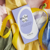 Ein lavendelfarbenes "mehr wir." - Kartenset von beherzt. ruht auf weichen pastellfarbenen Stoffen mit einem gelben Band und einer orangefarbenen Strickmöhre. Dieses Kartenset ermutigt zu offenen Gesprächen über Schwangerschaft, Geburt und eine selbstbestimmte Elternschaft.