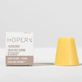 Eine weiß-beige Schachtel mit der Aufschrift Hopery Ultra Rich Face Serum Normal Skin steht neben einem gelben, kegelförmigen, festen Gesichtsserum mit Bio-Arganöl auf einer hellen Fläche vor einem unifarbenen Hintergrund.