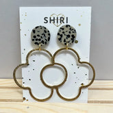 Ein Paar Shiri Ohrringe "Casablanca" mit schwarz-weiß gepunkteten Nieten und großen goldenen blumenförmigen Reifen - ein auffälliger Statement-Schmuck von Schmuck aus Hannover - präsentiert auf einer weißen Karte mit SHIRI und einem goldenen Herz.