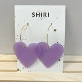 Ein Paar große, lilafarbene Ohrringe in Herzform "Havanna" von Shiri mit dünnen Goldreifen wird auf einer weißen SHIRI-Karte vor hellem Holz und einer grauen Wand präsentiert und verleiht jedem Look einen lebendigen Charme.