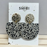 Ein Paar Shiri Ohrringe "Kairo" mit runden, strukturierten Goldnieten und großen, schwarz-weiß gesprenkelten Halbmond-Anhängern - Statement-Schmuck von Shiri, präsentiert auf weißer Karte mit SHIRI-Logo und Herz.