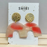 Ein Paar Shiri Ohrringe "Las Vegas" mit strukturierten goldfarbenen runden Nieten und durchscheinenden beigefarbenen und korallenfarbenen gewölbten Anhängern, präsentiert auf einer weißen SHIRI-Karte mit Herz als Statement-Schmuck aus Hannover.