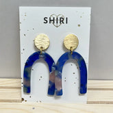 Ein Paar Schiri-Ohrringe "Los Angeles" mit goldenen Rundnieten und blau marmorierten Bogenanhängern wird auf einer weißen SHIRI-Karte mit Herzumriss präsentiert. Ideal für Statement-Schmuck Hannover Fans.