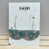 Ein Paar Ohrringe "Nizza" von Shiri mit großen goldenen Reifen und blau-weiß marmorierten Acetat-Anhängern, die auf einer Markenkarte mit einem goldenen Herz über dem Namen präsentiert werden.