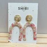 Die Ohrringe "Orlando" von Shiri bestehen aus strukturierten, runden goldenen Ohrsteckern und U-förmigen Acetat-Ohrringen in transluzentem Rosa und Weiß, die auf einer Shiri-Markenkarte präsentiert werden. Schmuck aus Hannover.