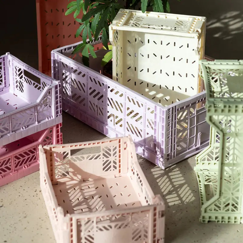 Pastellfarbene Aykasa Ay-Kasa Klappbox Midi Kisten in Rosa, Lavendel, Creme und Mintgrün sind auf einer sonnenbeschienenen Fläche gestapelt, im Hintergrund ist eine grüne Pflanze zu sehen.