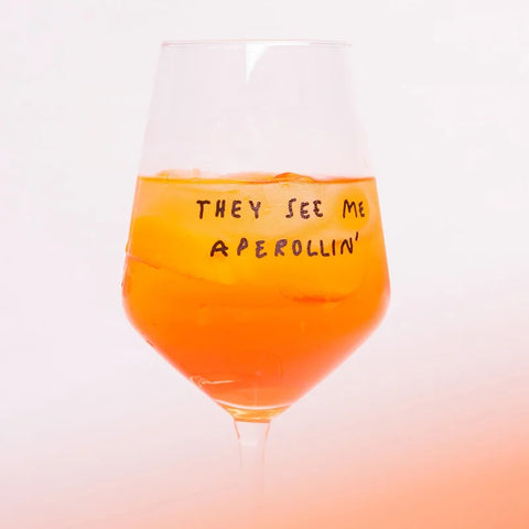 Ein Glas Orangen-Aperitif mit Eis steht in dem Weinglas "They See Me Aperollin'" von Johanna Schwarzer für Selekkt, das mit seinem verspielten handgeschriebenen Text vor einem zartrosa Hintergrund aufwartet.
