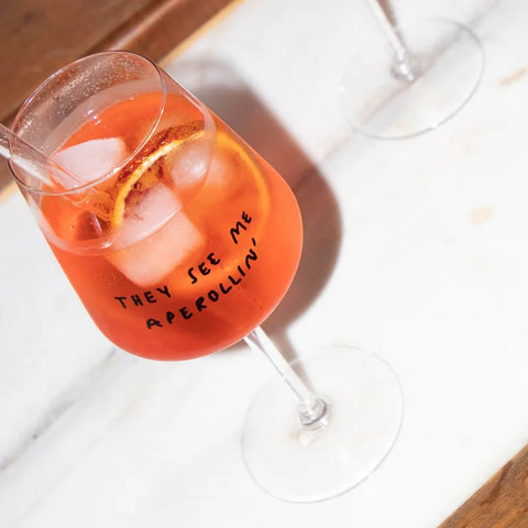 Ein "They See Me Aperollin'" Weinglas von Johanna Schwarzer (Selekkt), spülmaschinengeeignet, gefüllt mit orangefarbenem Aperol Spritz, Eis und einer Orangenscheibe ruht auf einer weißen Fläche.