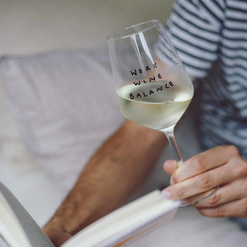 Eine Person im gestreiften Hemd hält das Weinglas "Work Wine Balance" von Johanna Schwarzer von Selekkt mit Siebdruck-Schriftzug in der Hand, während sie ein Buch liest.