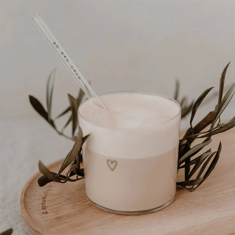 Ein Glas mit Kaffeeschaum und Eulenschnitt Strohhalme "No Bad Days" schwarz aus Glas 20cm (4er Set) steht auf einem Holztablett mit Olivenzweigen. Die minimale Szene ist sanft beleuchtet, und das Glas zeigt ein kleines Herzmotiv.