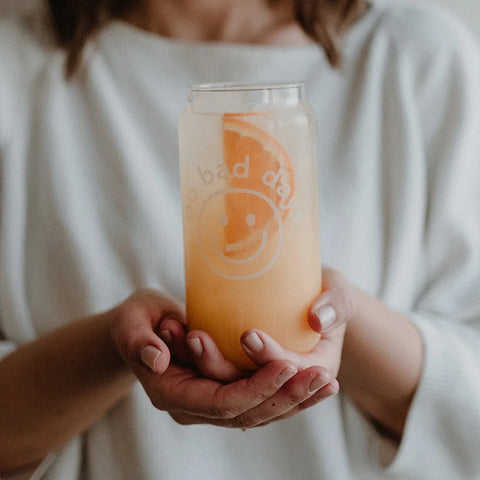 Eine Person in einem weißen Oberteil hält das Eulenschnitt Hohes Trinkglas "No Bad Days", ein klares Glas mit einem Smiley-Gesicht und einem fröhlichen Text, gefüllt mit Orangensaft, einem Eiswürfel und einer Grapefruit-Scheibe.