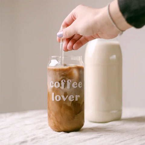 Eine Hand rührt Eiskaffee mit einem Glasstrohhalm im Eulenschnitt Hohes Trinkglas "Coffee Lover" um. Eine Flasche Milch im Hintergrund vervollständigt diese leichte Szenerie, ideal für den Genuss von Sommercocktails.