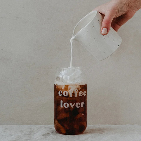 Eine Hand gießt Milch aus einer weißen Tasse in ein Eulenschnitt Hohes Trinkglas "Coffee Lover" aus Borosilikatglas, das mit Eiskaffee gefüllt ist und den Schriftzug "coffee lover" trägt.