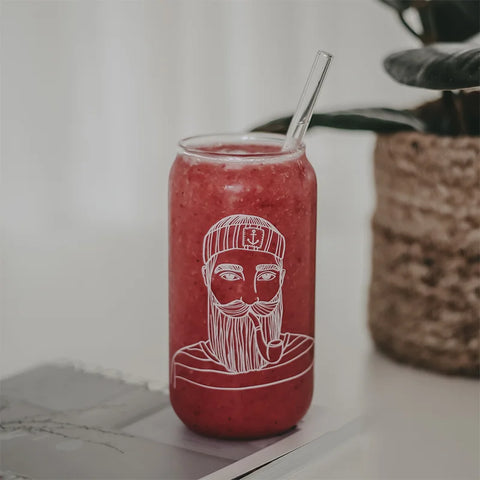 Ein leuchtend roter Smoothie mit einem durchsichtigen Strohhalm steht auf einem Magazin im Hohes Trinkglas "Seemann" von Eulenschnitt, das eine weiße Strichzeichnung eines bärtigen Mannes mit einer Mütze zeigt. Eine Topfpflanze steht unscharf im Hintergrund.