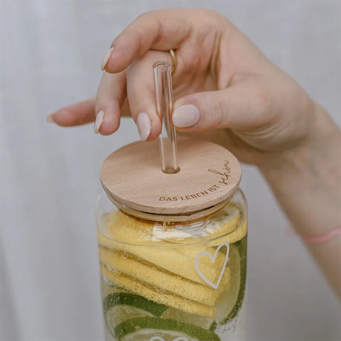 Eine Hand hält ein Glas mit Zitronen- und Limettenscheiben, darauf der Eulenschnitt-Deckel "Das Leben ist schön" für Hohes Trinkglas aus FSC-zertifiziertem Buchenholz, mit klarem Strohhalm und eingraviertem Text. Ein weißes Herz ziert das Glas.