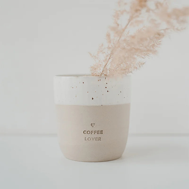 Der Becher "Coffee Lover" aus Steingut von Eulenschnitt in Beige und Weiß zeigt geprägten Schriftzug und präsentiert filigranes, getrocknetes Gras vor einem minimalistischen, hellen Hintergrund.
