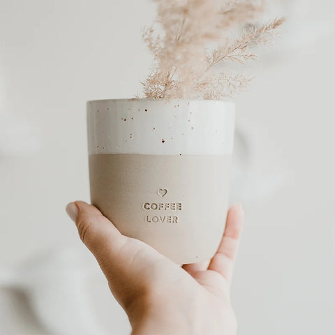 Eine Hand hält den Eulenschnitt Becher "Coffee Lover" aus Steingut, geprägt mit COFFEE LOVER und gestylt mit hellbraunem getrocknetem Gras vor einem sanft verschwommenen Hintergrund.