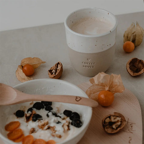 Eine Schale Joghurt mit Blaubeeren, Walnüssen und Orangenfrüchten steht neben einem Holzlöffel. Daneben steht der Eulenschnitt Becher "Coffee Lover" aus Steingut, der neben Walnüssen und Orangenbeeren für Gemütlichkeit sorgt.
