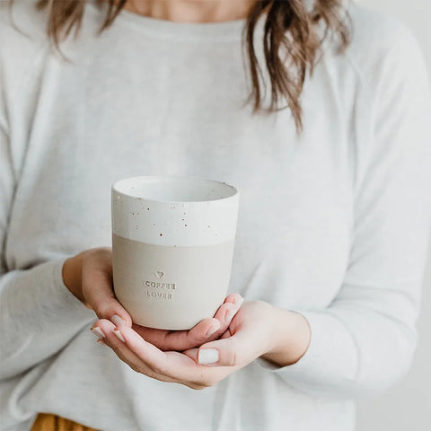 Eine Frau in einem hellgrauen Oberteil hält den Eulenschnitt Becher "Coffee Lover" aus Steingut, einen beigen Keramikbecher mit COFFEE LOVER und einem kleinen geprägten Herz. Nur ihre Hände und ihr Oberkörper sind zu sehen.