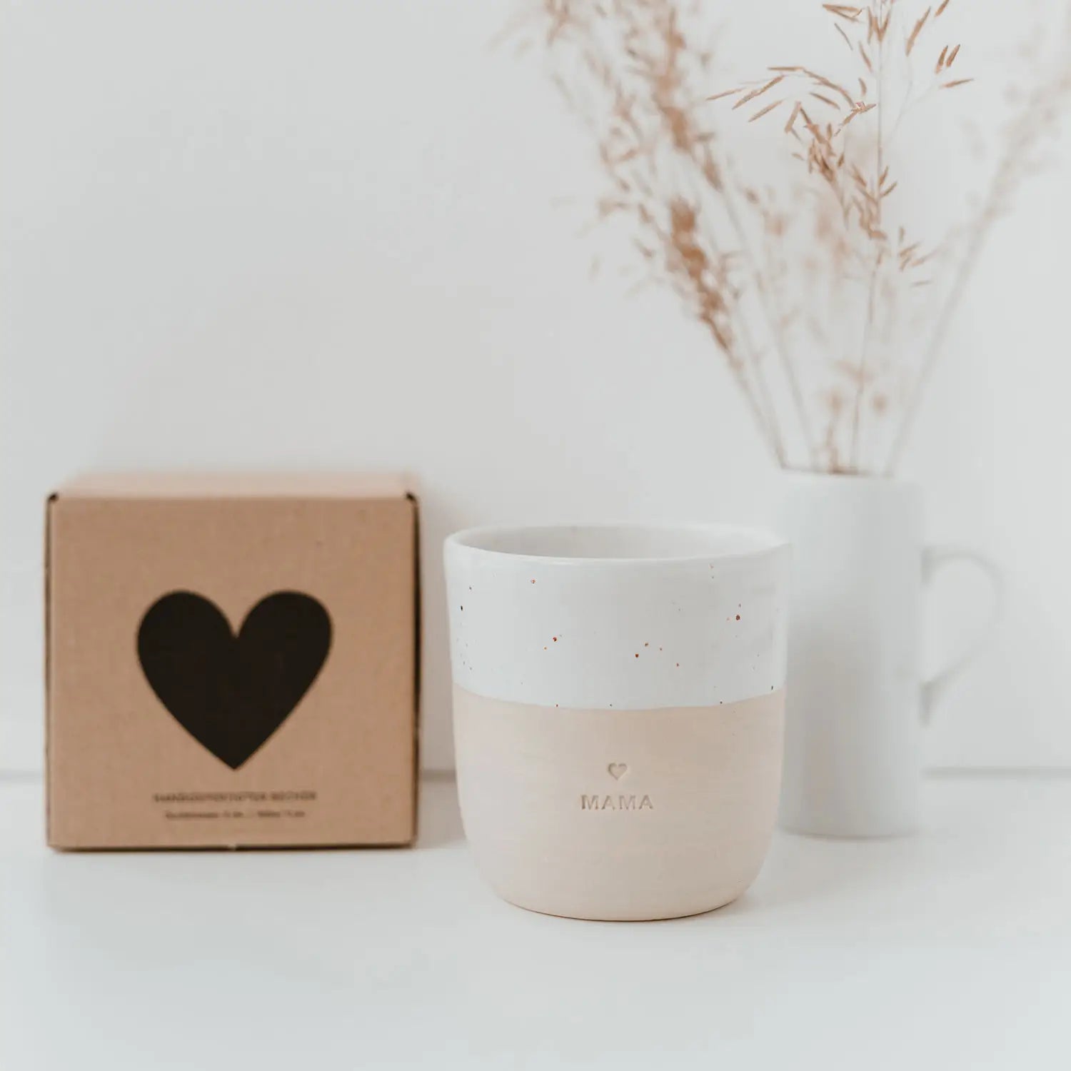 Der Becher "Mama" aus Steingut von Eulenschnitt in Beige-Weiß steht auf einer weißen Fläche neben einer braunen Herz-Box. Im Hintergrund eine Vase mit Trockenblumen - ein ideales Geschenk für Mami im sanften, minimalistischen Stil.