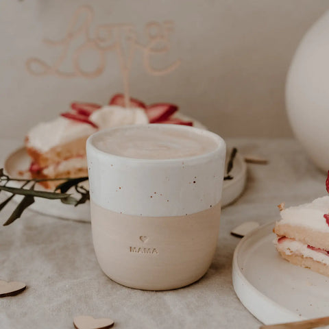 Der Becher "Mama" aus Steingut von Eulenschnitt steht mit eingraviertem Herz auf einem Tisch neben Tellern mit Erdbeerkuchen und einem "Love"-Kind - das ideale Geschenk in sanft neutraler Ästhetik für jede Mama.