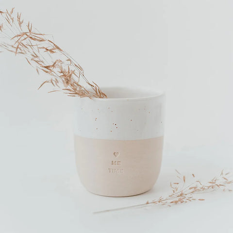 Der Eulenschnitt Becher "Me Time" aus Steingut, zweifarbig gestaltet und mit Herz sowie Schriftzug verziert, präsentiert zartes Trockengras vor einem minimalistischen, hellen Hintergrund.