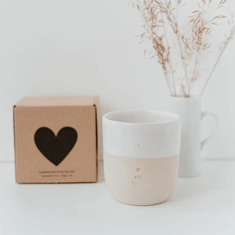 Der Becher "Me Time" aus Steingut von Eulenschnitt steht auf weißem Untergrund, davor eine braune Box mit Herz-Ausschnitt und eine weiße Vase mit Trockenblumen. Der handgemachte Becher wurde in Portugal gefertigt.