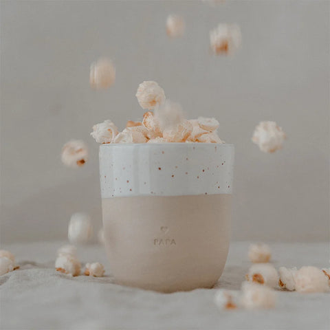 Der beige Becher "Papa" aus Steingut von Eulenschnitt, gefüllt mit Popcorn und überquellenden Körnern, stilvoll vor neutralem Hintergrund. Handgemacht in Portugal für einzigartige Genussmomente.