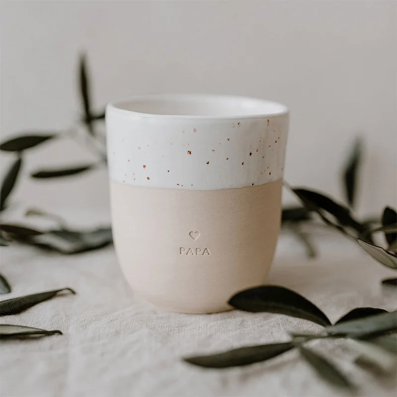 Der Becher "Papa" aus Steingut von Eulenschnitt ist handgemacht, hat einen weiß gesprenkelten und beigen Look, ein geprägtes Herz sowie den Schriftzug PAPA. Grüne Blätter umgeben den Becher auf hellem Stoff.