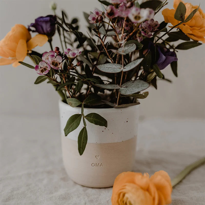 Der Becher "Oma" aus Steingut von Eulenschnitt präsentiert einen bunten Strauß aus orangefarbenen Ranunkeln, lila und kleinen rosa Blüten mit grünen Blättern - ein liebevolles Geschenk für Oma. Eine orangefarbene Blume liegt daneben auf weißem Stoff.