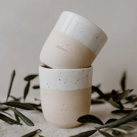 Zwei Eulenschnitt-Becher "Oma" aus Steingut, im weiß-beigen Design gestapelt und von grünen Blättern umgeben - ein liebevolles Geschenk für Oma.