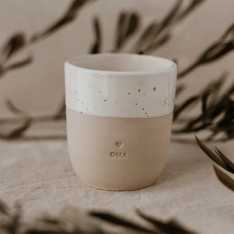 Der Becher "Oma" aus Steingut von Eulenschnitt in Beige mit weiß gesprenkelter Oberhälfte und Herzgravur steht auf hellem Stoff mit Pflanzenschatten - ein besonderes Geschenk für Oma.