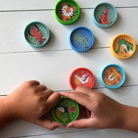 Ein Kind hält Rex Londons Geduldsspiel "Bunte Kreaturen", ein kleines rundes Labyrinthspielzeug, das zusammen mit anderen lebendigen Tiermotiven auf einer weißen Holzoberfläche gezeigt wird.