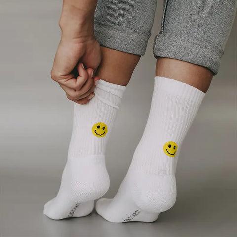 Eine Person in hochgekrempelten grauen Hosen passt ihre Eulenschnitt Socken "Smiley" gelb an, die auf der Rückseite in der Nähe des Knöchels einen gelben Smiley haben. Der Hintergrund ist einfarbig und grau.
