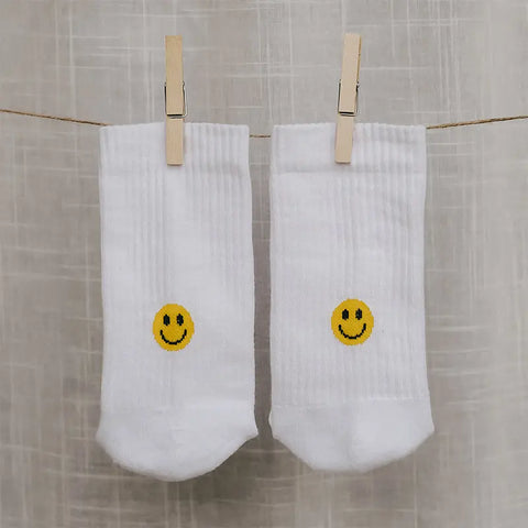 Zwei Socken "Smiley" gelb von Eulenschnitt aus weißer Bio-Baumwolle hängen mit Holzklammern an einer Wäscheleine vor hellem, strukturiertem Hintergrund.