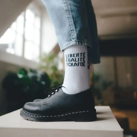 Eine Person in hochgekrempelten Jeans und schwarzen Schnürschuhen trägt die FCKAFDÉ Statement Socken - weiß von Arrel mit dem Schriftzug LIBERTE EGALITE FCKAFDE und steht in einem Innenraum auf einem hellen Podest mit einem leicht verschwommenen Hintergrund.