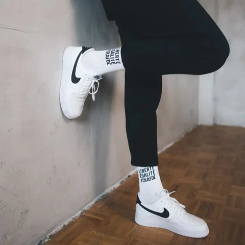Person trägt schwarze Hose, weiße Nike-Sneaker und Arrel FCKAFDÉ Statement Socken - weiß aus Bio-Baumwolle mit LIBERTE EGALITE FRATRITE und FCKAFD-Aufschrift und steht an heller Wand auf Holzboden.