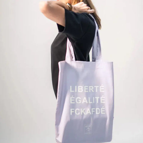 Eine Person in einem schwarzen Hemd trägt die Arrell FCKAFDÉ Premium Tote Bag - Purple & Vanilla Ice aus Bio-Baumwolle mit weißem LIBERTÉ ÉGALITÉ FCKAFDÉ-Schriftzug. Der Hintergrund ist einfarbig und hell gefärbt.
