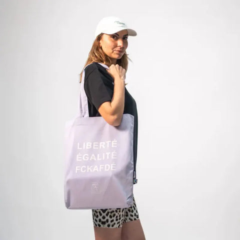 Eine Frau mit weißer Mütze, schwarzem T-Shirt und Shorts mit Leopardenmuster steht seitlich und hält die Arrell FCKAFDÉ Premium Tote Bag - Purple & Vanilla Ice mit dem fettgedruckten weißen Text LIBERTÉ ÉGALITÉ FCKAFDÉ vor einem schlichten hellen Hintergrund.