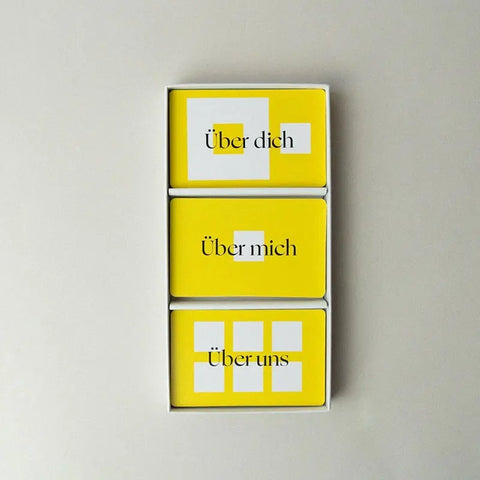Eine Schachtel der "90 Fragen, die verbinden." - Kartenset von beherzt. mit drei gelben Karten - über dich, über mich, über uns - in einer Reihe auf hellem Hintergrund. Jede Karte zeigt weiße und gelbe Formen für mutige Gespräche mit 90 Fragen.
