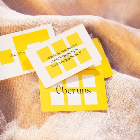Drei gelb-weiße Gesprächskarten aus dem "90 Fragen, die verbinden."-Kartenset von beherzt. mit deutschem Text liegen auf beigem Stoff. Eine Karte fragt: Was ist dir von unserer ersten Begegnung in Erinnerung geblieben?.