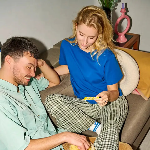 Eine lächelnde Frau in blauem Hemd und karierter Hose sitzt auf einer Couch und hält die "90 Fragen, die verbinden." - Kartenset von beherzt., während ein Mann in einem grünen Hemd in der Nähe sitzt, lächelnd und nach unten schauend. Im Hintergrund ist ein dekorierter Beistelltisch zu sehen.