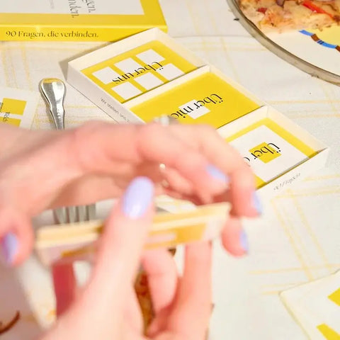 Eine Person mit hellviolettem Nagellack hält ein gelbes "90 Fragen, die verbinden." - Kartenset von beherzt. auf einem Tisch, der mit einer Gabel, Pizza und der Spieleschachtel gedeckt ist.