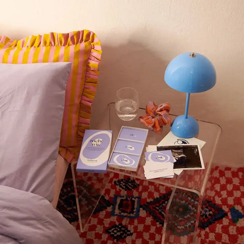 Auf einem durchsichtigen Nachttisch stehen eine blaue Lampe, ein Glas Wasser, ein Haargummi und mehrere pastellfarbene "mehr wir." - Kartenset-Pakete von beherzt. In der Nähe steht ein Bett mit gerüschten Kissen und rosa-gelben Streifen auf dem Bettvorleger.