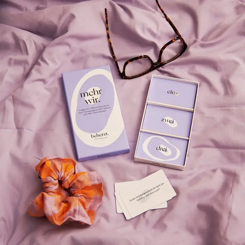 Ein lavendelfarbenes Kartenset mit dem Namen "mehr wir." von beherzt. liegt auf einer lila Tagesdecke mit einer Schildpattbrille und einem orangen Haargummi. Das Set enthält Karten mit der Aufschrift eins, zwei, drei und Fragen rund um die Schwangerschaft.