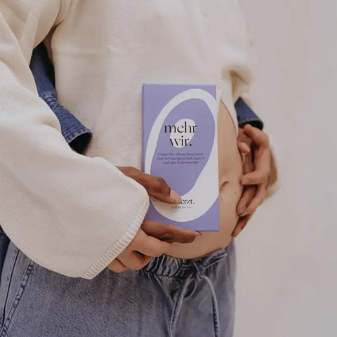 Jemand trägt einen leichten Pullover und Jeans und hält die "mehr wir." - Kartenset von beherzt. mit deutschem Text gegen ihren nackten Bauch, was auf eine Schwangerschaft oder Selbstfürsorge hindeutet. Der Fokus liegt auf der Karte und ihrer sanften Berührung.